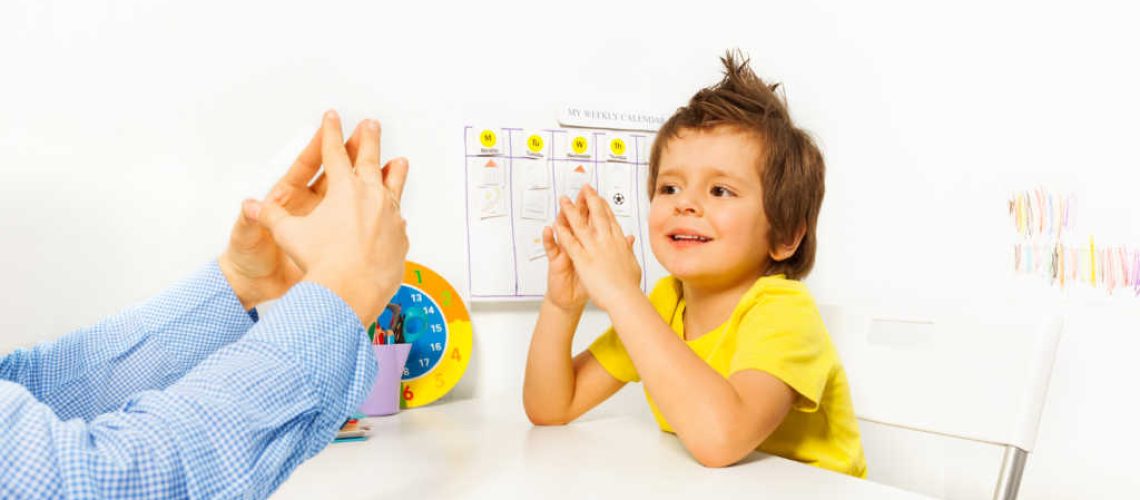 Smiling boy exercises putting hands and fingers together with therapist improving motor skills sitting opposite at the table indoors